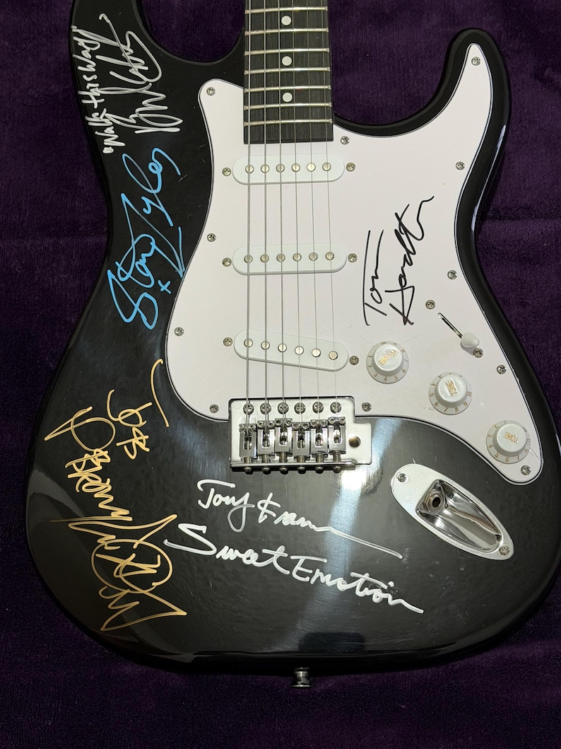 May include: Black electric guitar with white pickguard and silver hardware. The guitar features multiple signatures in various colors, including gold, blue, and white. The guitar has six strings and three white pickups.