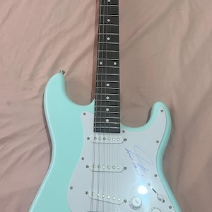 以下が含まれることがあります： 白のピックガードとシルバーのハードウェアを備えた、水色のエレキギター。ギターにはサインと「Good luck with the guitar. I hope you enjoy it.」の文字が入っています。