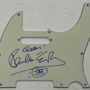 May include: Cream-coloured guitar pickguard with a blue ink signature and the word "cheers!". The pickguard features screw holes and a pickup cutout. A small drawing is below the signature.