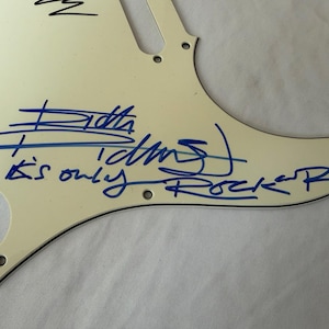 May include: Off-white guitar pickguard with blue handwritten text and signature. The text reads "Keith Richards" and "It's only Rock 'n Roll". The pickguard has several screw holes and a curved shape.