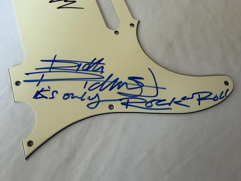 May include: Off-white guitar pickguard with blue handwritten text and signature. The text reads "Keith Richards" and "It's only Rock 'n Roll". The pickguard has several screw holes and a curved shape.