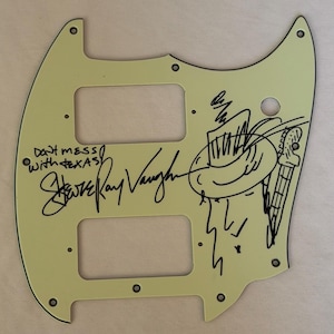 May include: A cream-colored guitar pickguard with black text and illustrations. The text reads "Don't mess with Texas" and "Stevie Ray Vaughan" in a signature style. A cartoon of a musician with a hat and guitar is also present.