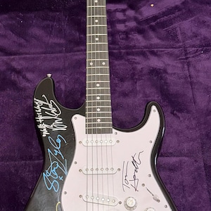 May include: Black electric guitar with a white pickguard and multiple signatures in various colors. The guitar has a wooden neck and headstock, with silver tuning pegs and a bridge. The body is glossy black, and the signatures appear to be from musicians.