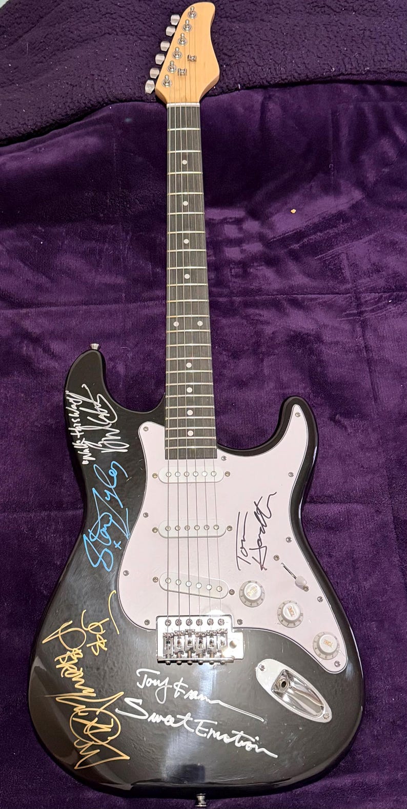 May include: Black electric guitar with a white pickguard and multiple signatures in various colors. The guitar has a wooden neck and headstock, with silver tuning pegs and a bridge. The body is glossy black, and the signatures appear to be from musicians.