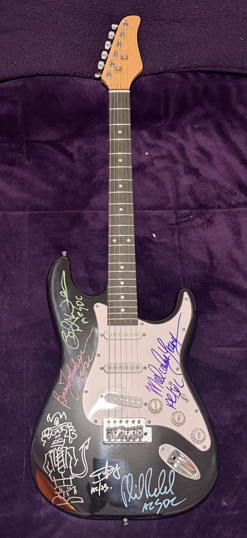 Pu&ograve; includere: Chitarra elettrica nera con manico e paletta in legno chiaro. La chitarra ha un battipenna bianco ed &egrave; ricoperta di firme. Il corpo della chitarra presenta un disegno a cartoni animati e la scritta "AC/DC".