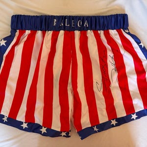 May include: Boxing shorts with a red and white striped design, blue trim, and white stars. The waistband is blue with the word "BALEGA" in white. A signature is visible on the shorts.