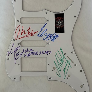 May include: White guitar pickguard with multiple signatures in various colors. A black and red Mot&ouml;rhead logo sticker is affixed to the upper right. Signatures include red, blue, purple, and green ink.