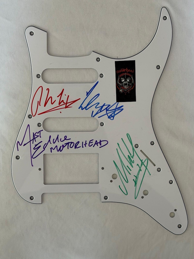 May include: White guitar pickguard with multiple signatures in various colors. A black and red Mot&ouml;rhead logo sticker is affixed to the upper right. Signatures include red, blue, purple, and green ink.