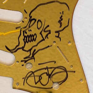 May include: A gold-colored guitar pickguard featuring a black skull illustration and handwritten signatures. The pickguard has several screw holes and a cut-out for the guitar's pickups. The skull design is detailed with sharp lines and shading.