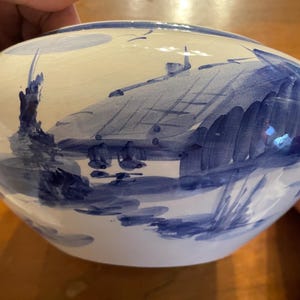 May include: A ceramic bowl with a hand-painted landscape scene in shades of blue. The design features a house, trees, and water, all rendered in a stylized, illustrative manner. The bowl has a glossy finish.