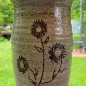 May include: A handmade ceramic vase with a light brown speckled finish. The vase features a floral design of three sunflowers etched in dark brown. The vase is cylindrical with a slightly flared top, perfect for displaying flowers.