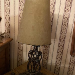 May include: A vintage table lamp with a beige lampshade and a decorative, dark metal base. The lampshade is conical and textured. The base features ornate, swirling designs. The lamp is on a wooden table.