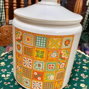 May include: White ceramic cookie jar with a lid. The jar is decorated with a colorful, geometric pattern of squares in shades of orange, yellow, green, and red. The jar is sitting on a green floral tablecloth.