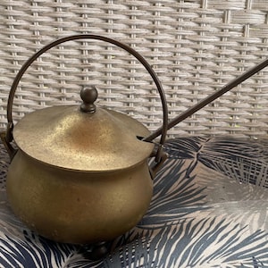 May include: A brass pot with a rounded body, a matching lid with a small knob, and three short feet. It features a curved swing handle and a long, slender metal rod extending from the side, designed to rest in a notch on the lid's edge.