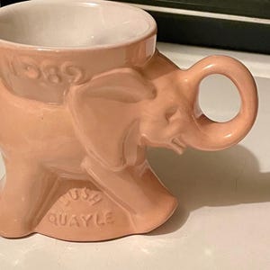 May include: A light pink ceramic mug shaped like an elephant with a trunk forming the handle. The mug features the embossed text 1989 near the top rim and the embossed text LUSH QUAYLE on the base. The interior of the mug is white.
