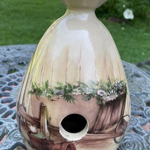 May include: A beige ceramic birdhouse with a decorative floral and wooden cart design. The birdhouse has a round opening and a flared top. The painted design includes a wooden cart filled with flowers and greenery.