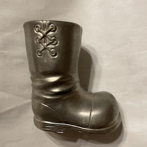 May include: A small, silver-colored boot figurine. The boot has a rounded toe, a ridged sole, and a decorative criss-cross pattern on the upper shaft. The boot is made of a solid, metallic material.
