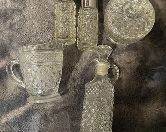 May include: A collection of clear glass serving pieces with a diamond pattern. The set includes two small shakers with silver lids, a creamer, a lidded jar, and a decanter with a handle.