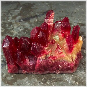 May include: A cluster of deep red crystals with a glittery texture. The crystals have a natural, jagged form and are set against a neutral background. The base of the cluster has a golden hue.