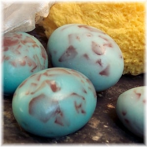 May include: Close-up of several oval-shaped soaps. The soaps are a light blue color with maroon-colored speckles. A yellow sponge and white mesh material are in the background. The soaps have a smooth, glossy finish.