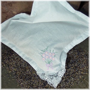 May include: A light blue, square-shaped sachet with a floral embroidery design. The embroidery features a bouquet of pink flowers with green leaves and a pink bow. The sachet has a white lace trim and is surrounded by dried lavender.
