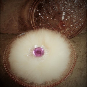 May include: A vintage pink glass powder jar with a fluffy white powder puff. The puff has a pink jewel in the center and sits inside the open jar. The lid is visible in the background.