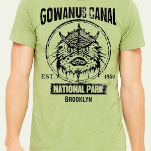 以下が含まれることがあります： 黒いモンスターのグラフィックプリントが施された緑色のTシャツで、"GOWANUS CANAL EST. 1860 NATIONAL PARK BROOKLYN"の文字が書かれています。