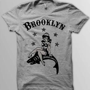 May include: A gray t-shirt with a black graphic of a pin-up girl sitting on a shark. The graphic also includes the word "BROOKLYN" and stars.