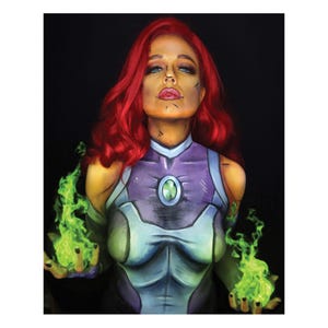 May include: A woman with red hair and green flames in her hands is painted to look like a comic book character. She is wearing a purple and green costume with a large green gem on her chest.