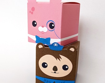 May include: Two small cardboard boxes with animal faces. The top box is pink with a bunny face, a blue bow tie, and a monocle. The bottom box is brown with a bear face and a blue sailor hat.