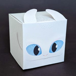 Könnte beinhalten: Eine weiße, quadratische Geschenkbox mit einem Cartoon-Drachengesicht, großen blauen Augen und kleinen weißen Hörnern. Die Box hat eine strukturierte Oberfläche und kleine Griffe oben.