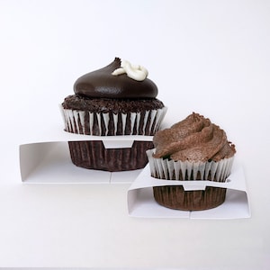 Puede incluir: Dos cupcakes de chocolate con glaseado blanco en la parte superior, exhibidos en soportes de cartón blanco para cupcakes.