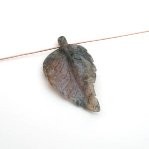 May include: A carved leaf pendant made of gray and green stone. The leaf has detailed veining and is suspended on a thin copper wire. The pendant is set against a white background.