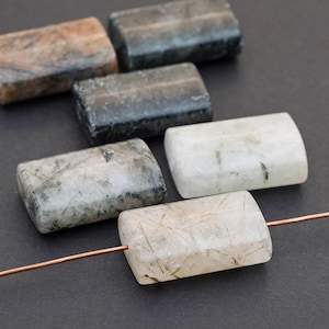 May include: A collection of rectangular gemstone beads in various colors, including gray, white, and brown. The beads are strung on a copper wire, showcasing their polished surfaces and natural inclusions. The beads are ideal for jewelry making.