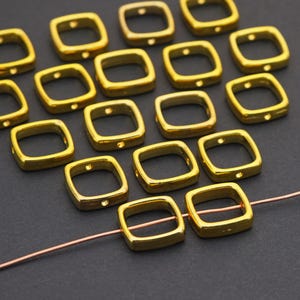 May include: A collection of small, square-shaped gold-colored metal beads with rounded corners and a central opening. Each bead features a single small hole on one side. Two of these beads are threaded onto a thin, straight copper-colored wire.