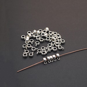 May include: A collection of small, silver-colored metal beads with a loop on one side. Some beads are strung on a thin copper wire. The beads are scattered on a dark surface, suggesting their use in jewelry making or crafts.