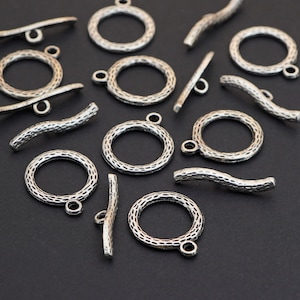 May include: A collection of silver-toned toggle clasps and ring clasps, scattered on a dark surface. The clasps have a textured, hammered appearance. The toggle clasps are bar-shaped, and the ring clasps are circular with a small loop.