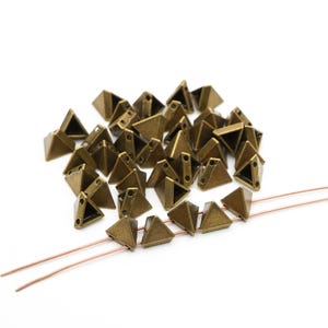 May include: A collection of bronze-colored, pyramid-shaped beads with two holes for stringing. The beads are scattered on a white surface, with two copper-colored wires visible. The beads have a metallic finish and a geometric design.