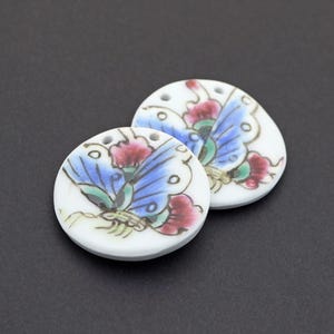 May include: Two circular white ceramic pendants featuring a hand-painted butterfly design. Each butterfly has blue wings with black outlines, red accents, and green details. The pendants have two small holes at the top for threading or attachment.