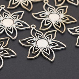 May include: Close-up of several silver-colored flower-shaped pendants. Each pendant has a detailed openwork design with a central circular area. The pendants are scattered on a dark surface, showcasing their intricate floral pattern.