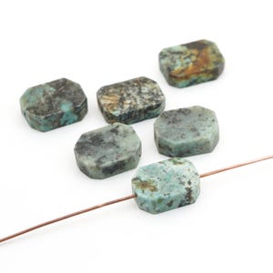May include: Six flat, rectangular turquoise beads with rounded corners, featuring natural dark brown and black matrix inclusions. One bead is threaded onto a thin copper-colored wire, while the other five are arranged loosely nearby.