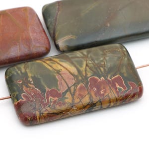 May include: Three rectangular, polished jasper beads featuring a smooth, flat surface. The stones display a marbled pattern with earthy tones of olive green, deep red, and golden brown, threaded on a thin copper wire through the center.