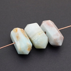 May include: Three faceted gemstone beads in shades of blue, white, and tan are strung on a copper wire. The beads have a polished finish and a geometric shape. The background is a solid black.