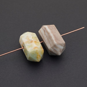 May include: Two faceted gemstone beads. One is a pale green with yellow and orange accents, the other is a light brown with darker brown stripes. Both are strung on a copper wire against a dark background. These beads are suitable for jewelry making.