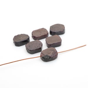 May include: Six dark reddish-brown, irregularly shaped, flat rectangular beads with smooth surfaces. One bead is threaded onto a thin, flexible copper-colored wire, while the other five beads are arranged in a cluster nearby on a white surface.