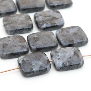 May include: A collection of square-shaped labradorite beads with a smooth, polished finish. The stones feature a dark gray base color with iridescent blue and silver inclusions throughout. Two of the beads are threaded onto a thin copper wire.