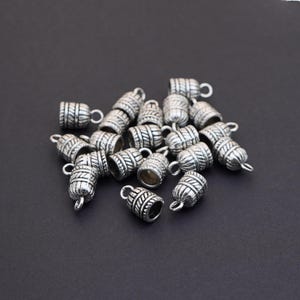 May include: A collection of silver-colored barrel-shaped jewelry beads with intricate etched designs. Each bead has a small loop at the top for stringing. The beads are scattered on a dark surface.