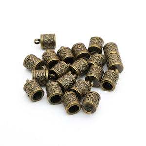 May include: A collection of antique bronze-colored barrel beads with intricate scrollwork patterns. Each bead has a small loop at the top for stringing. The beads are scattered on a white surface, showcasing their detailed design.