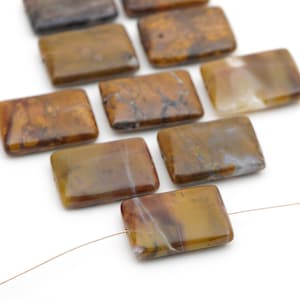 May include: A collection of rectangular, polished gemstone beads in shades of brown, tan, and cream. Each bead features unique, natural swirling patterns and inclusions. The beads are strung on a thin, metallic-colored wire, showcasing their smooth, flat surfaces and rounded edges.