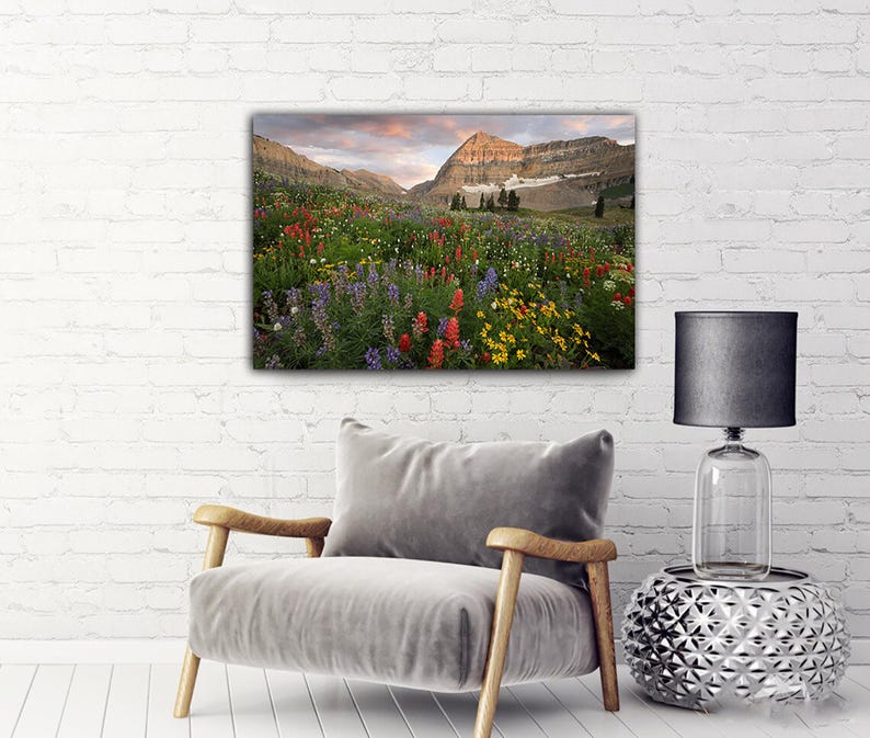 a large, colorful painting of a mountain landscape hanging on a white brick wall above a gray armchair in a living room setting.